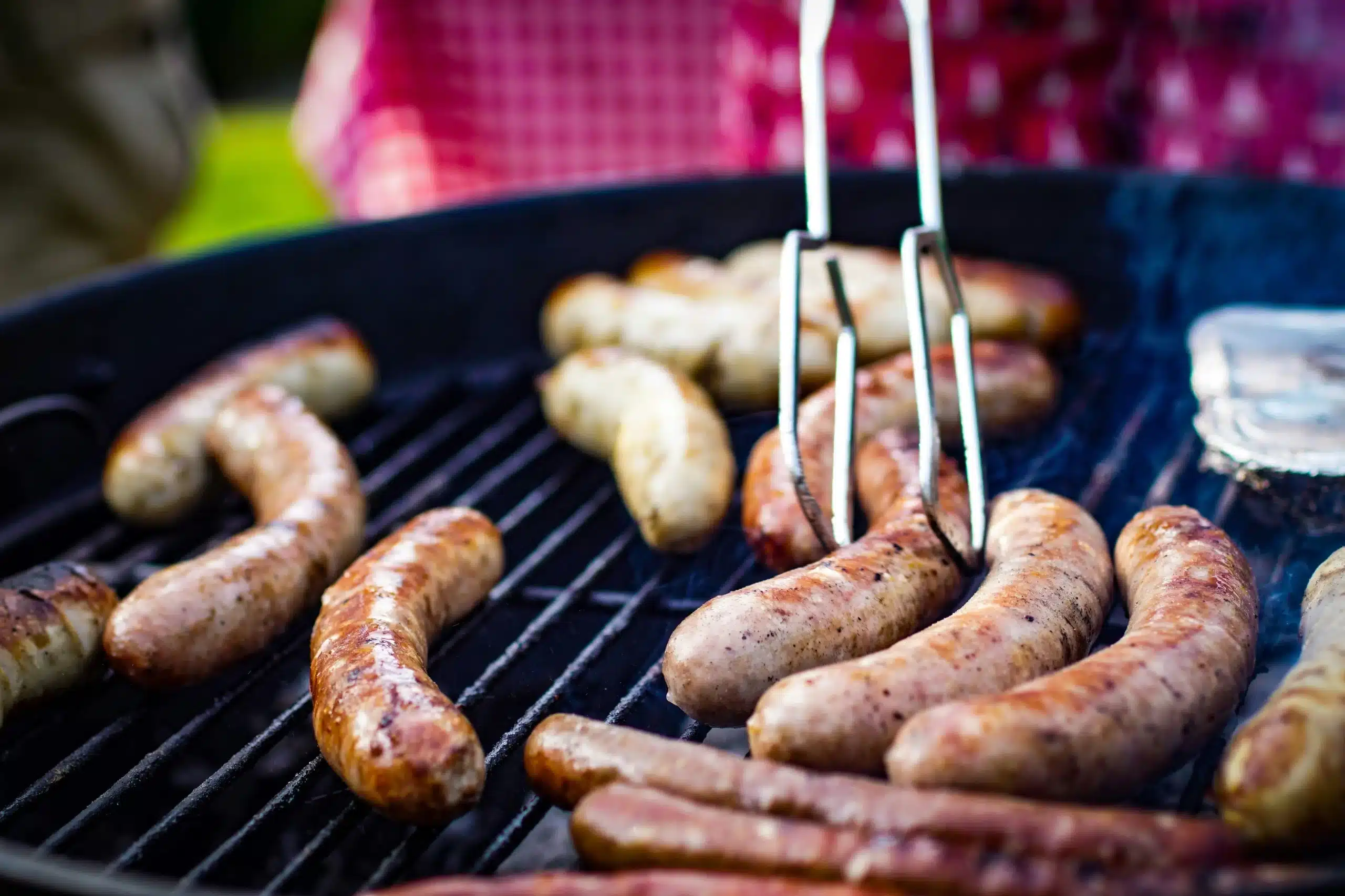 Sausages cooking on a charcoal grill, with tongs held above them and a red checkered tablecloth in the background, potentially highlighting the link between processed meats and cancer risks.