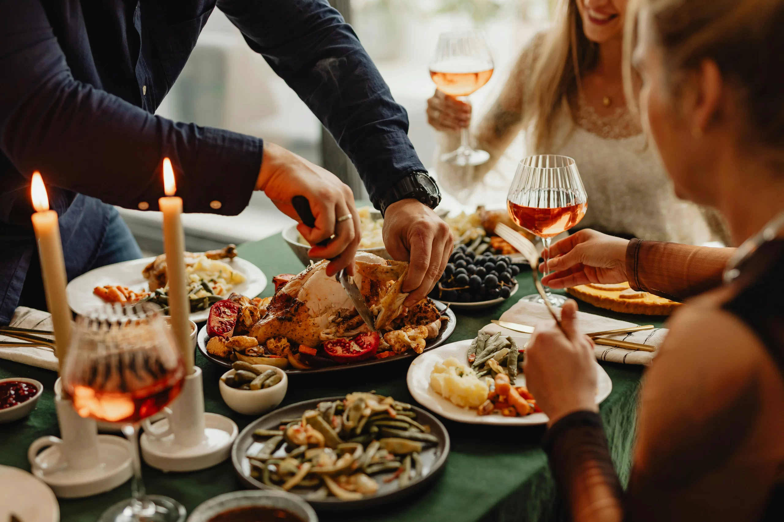 People enjoying a meal at a table set with candles, drinks, and dishes of vegetables and meats. A person is carving roasted meat while others hold glasses of wine.