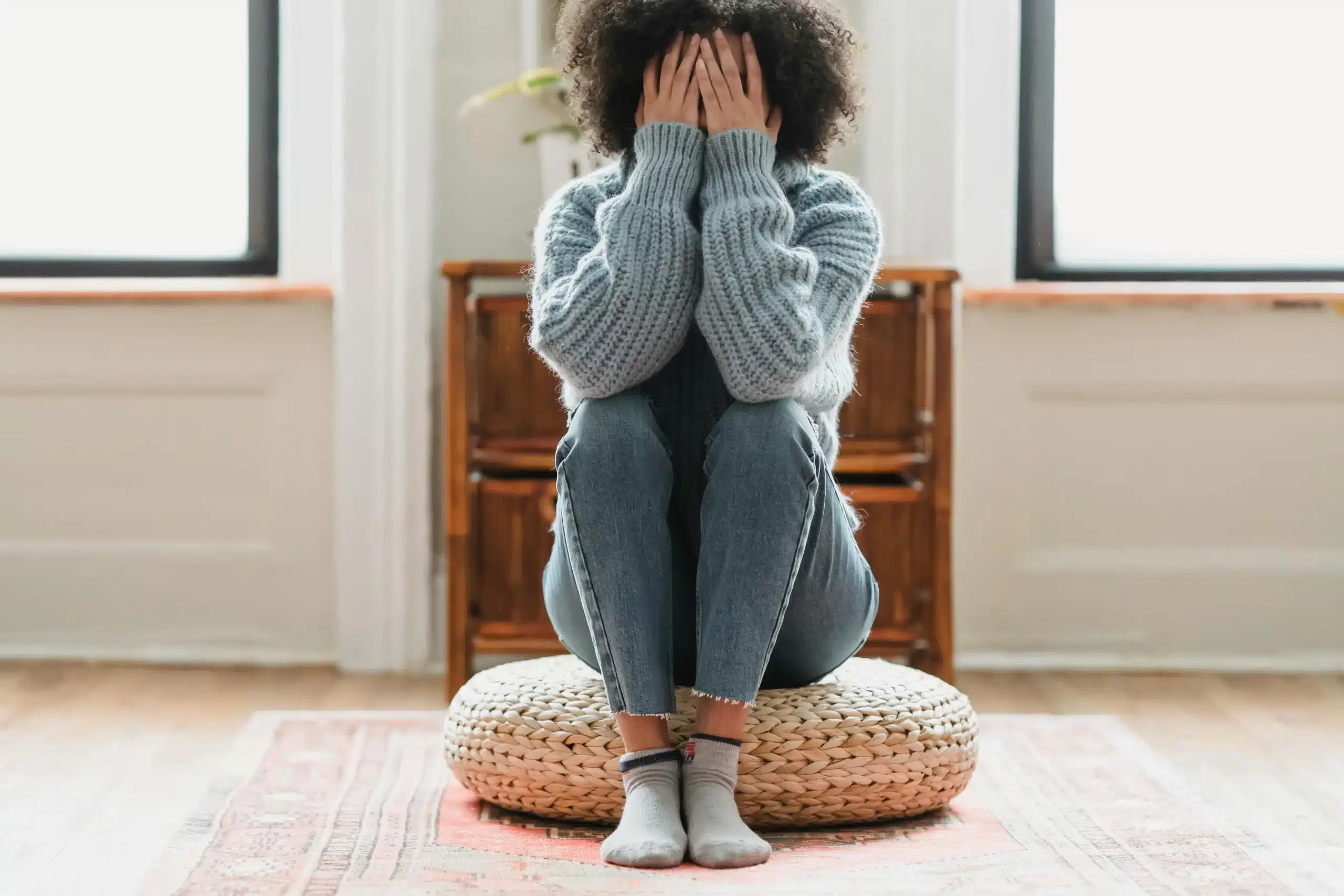 A person sitting on a woven pouf, covering their face with their hands, in a room with wooden floors and a large window, hoping to regulate cortisol levels.