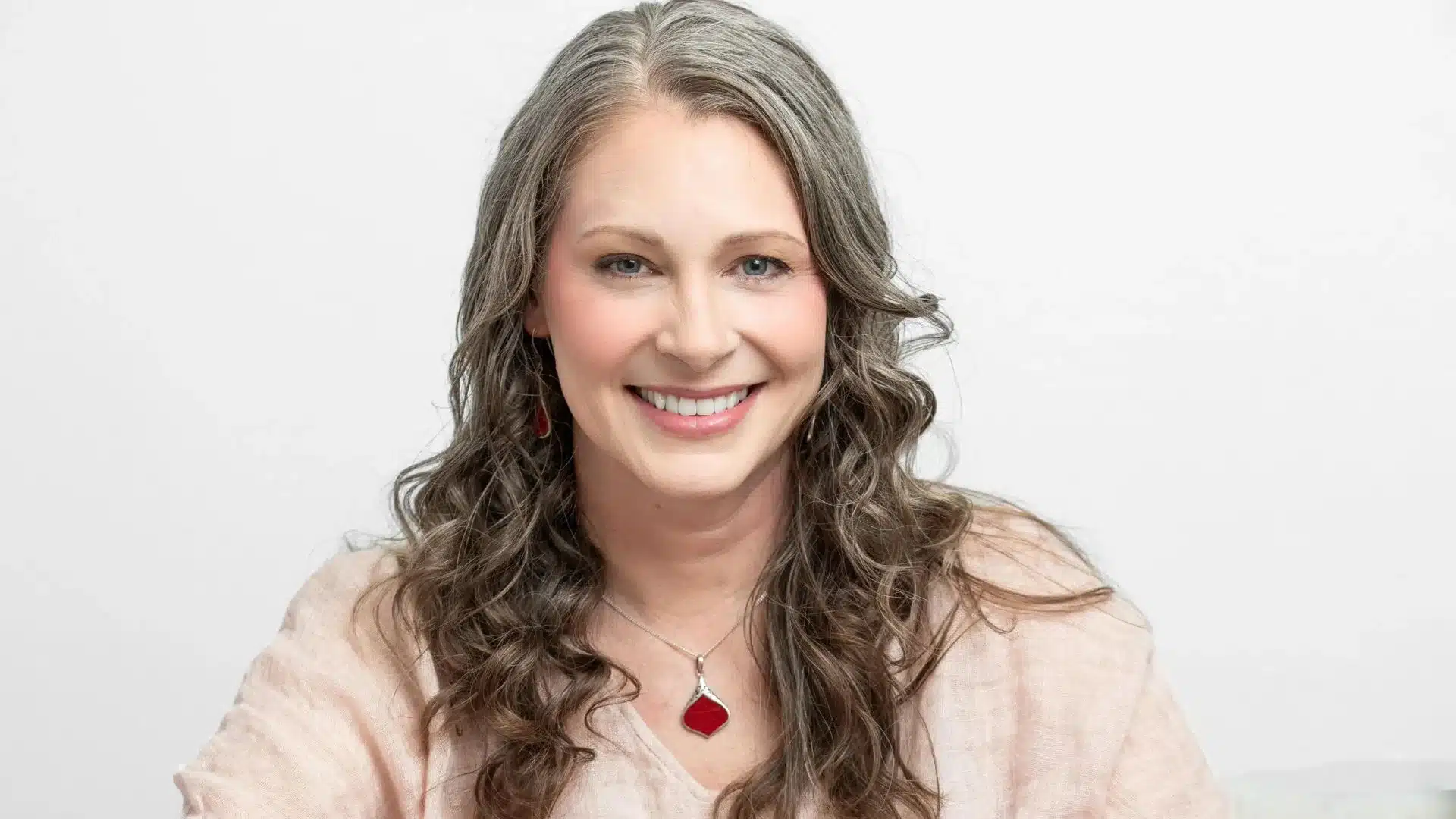 A person with long, wavy gray hair smiles, wearing a beige top and a red pendant necklace that symbolizes inner peace. They are set against a plain white background.