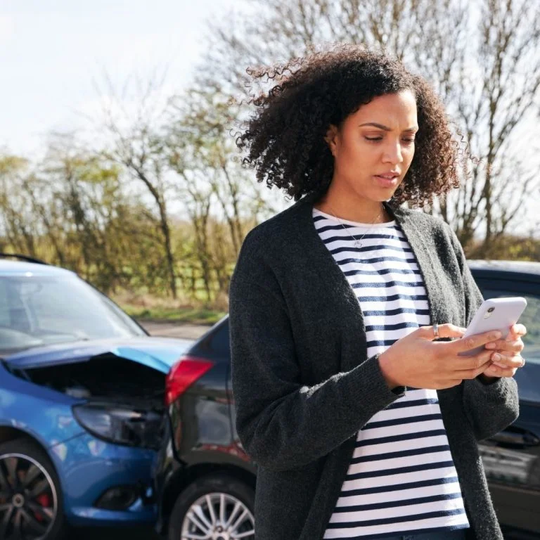 Woman looking at her phone in distress with a car accident behind her.