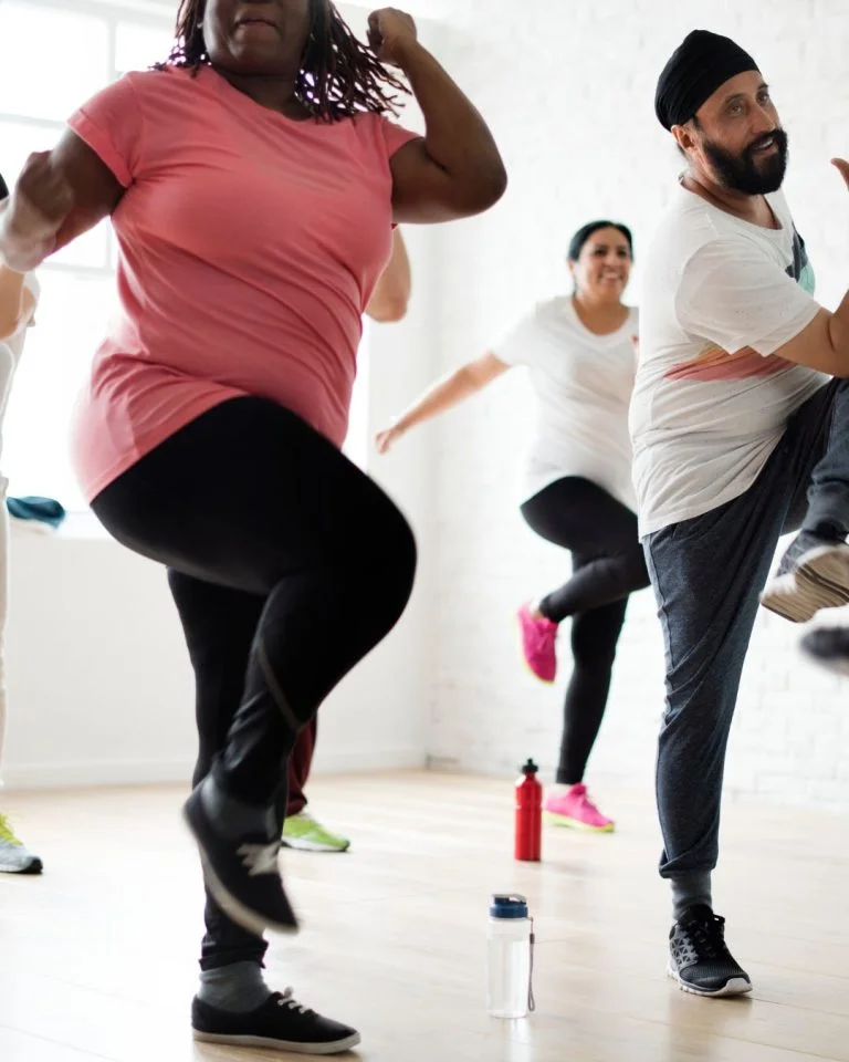 A diverse group of people taking part in a group fitness class in a bright, sunlit room with wooden floors. In the foreground, a Black woman in a pink t-shirt and black leggings and a man wearing a white shirt and black turban lift their knees high in sync.