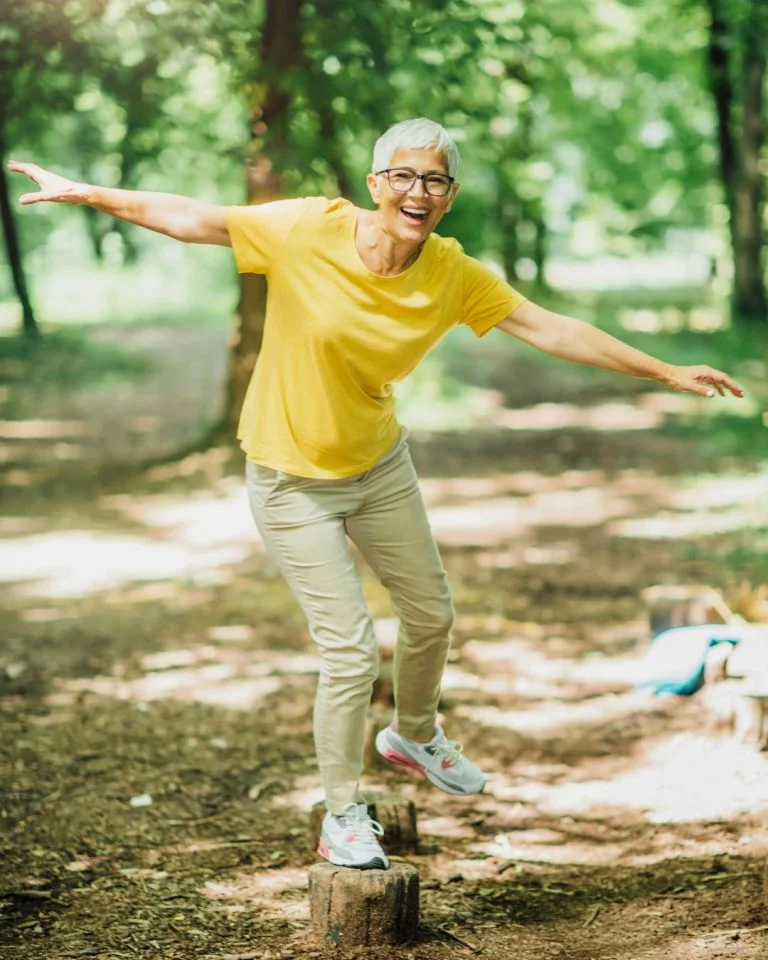 A smiling older woman with short white hair balancing on one foot on a wooden log in a sunny forest path.