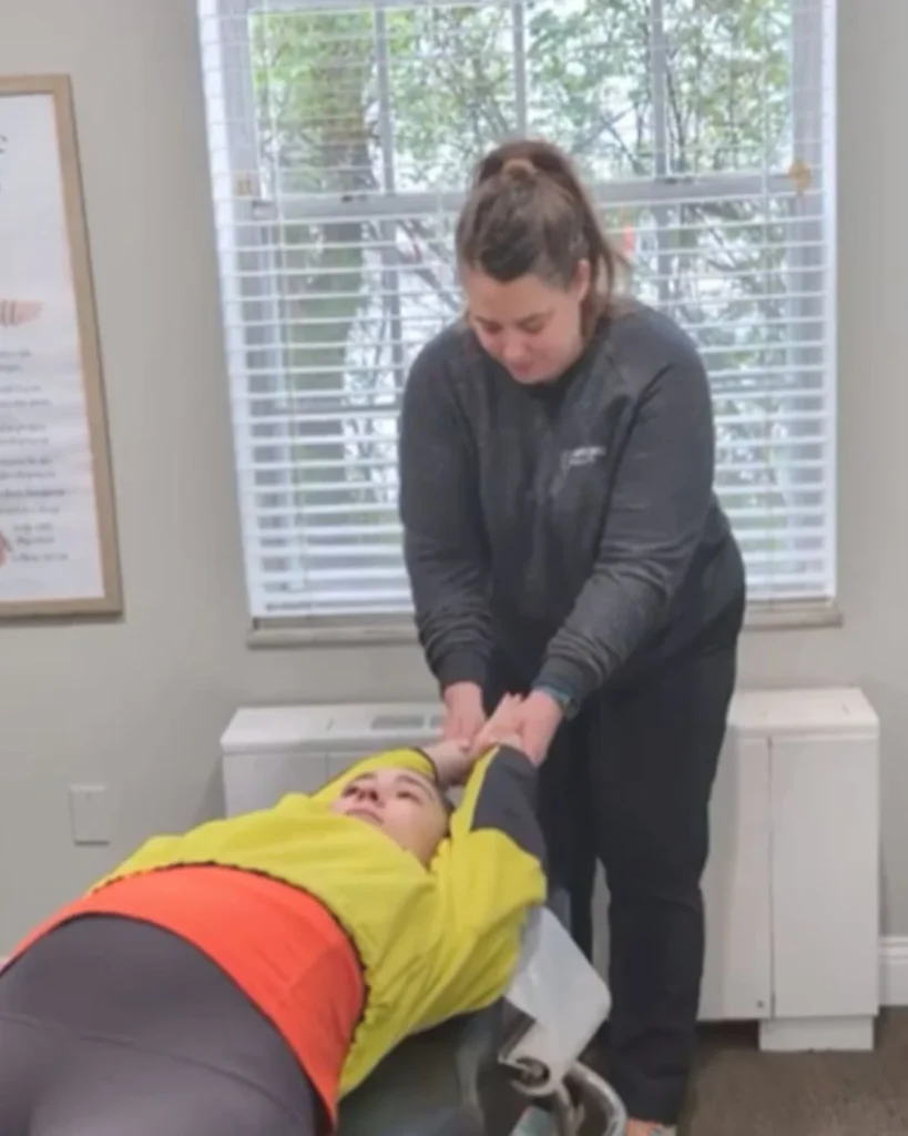 A female chiropractor holding the wrists of a patient lying face-up on an adjustment table to perform an upper body or shoulder stretch.