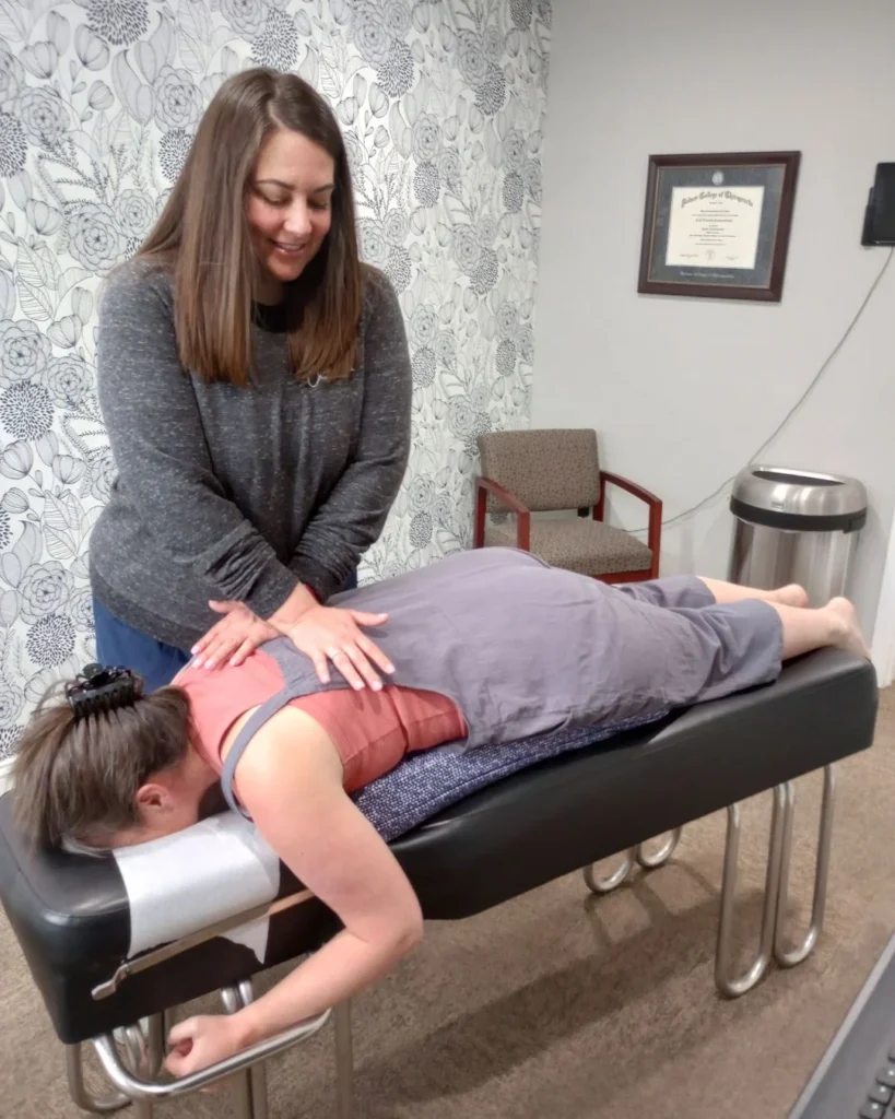 A female chiropractor gently placing her hands on the upper back of a pregnant patient lying face-down on a specialized pregnancy chiropractic cushion.