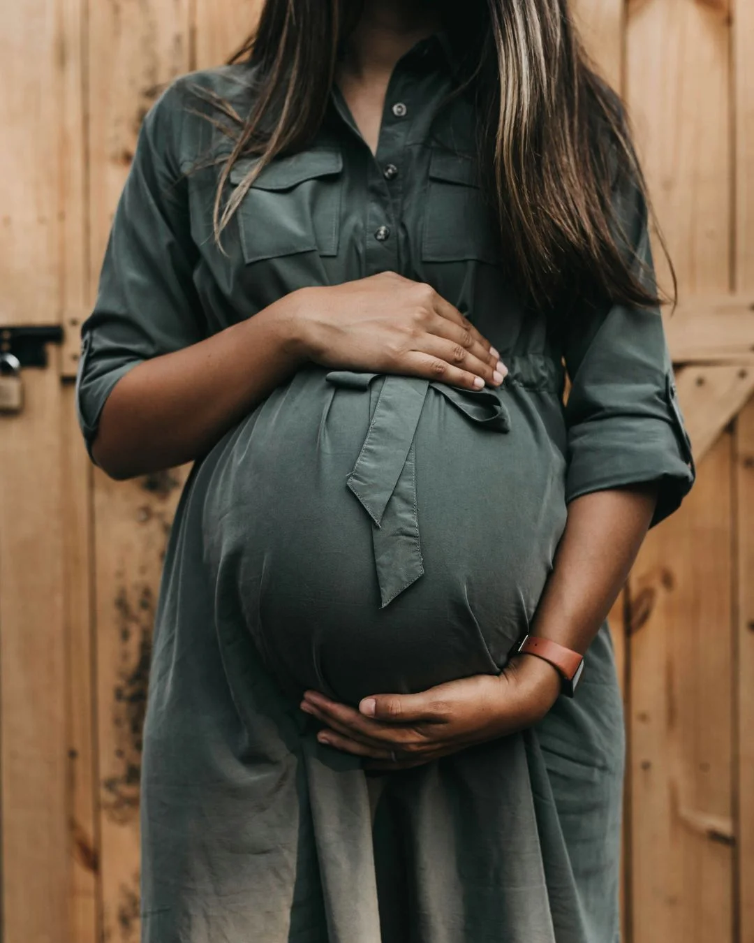 Midsection view of a pregnant woman in a dark green dress cradling her baby bump with both hands in front of a wooden background.