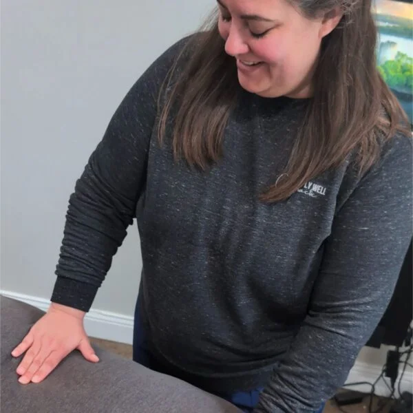 Smiling female chiropractor leaning over an adjustment table in a clinic, wearing a dark Simply Well Chiropractic sweatshirt.
