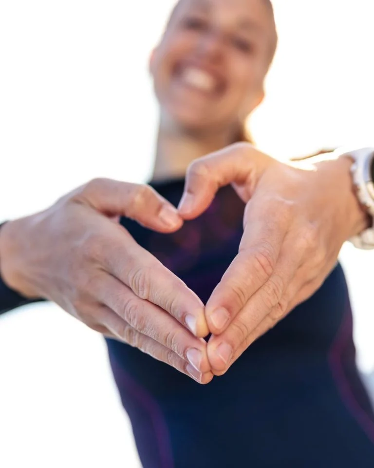 Close-up of a smiling woman in athletic gear making a heart shape with her hands toward the camera.