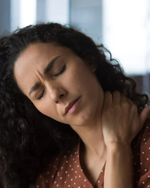 Close-up of a woman with dark curly hair wincing in discomfort while holding the back of her neck.