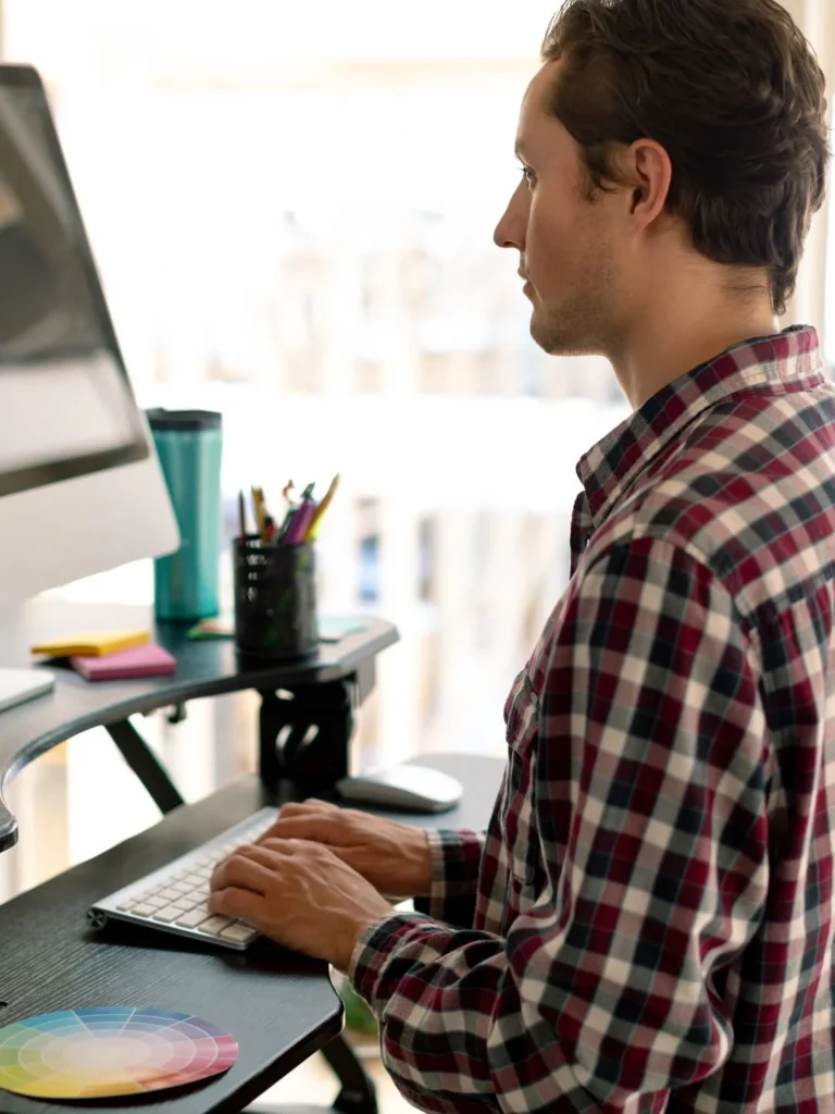 Side profile shot of a man wearing a red plaid shirt standing upright while typing on a keyboard at an adjustable standing desk.